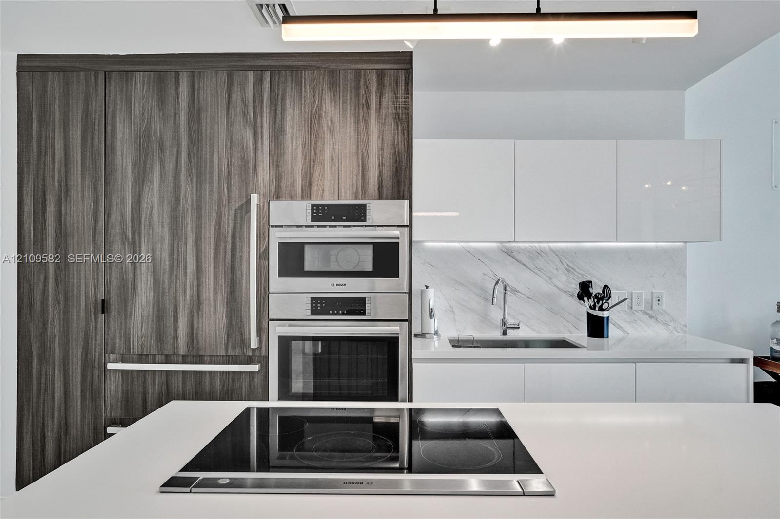 Kitchen featuring wood-finish floor-to-ceiling cabinetry, stainless steel built-in double ovens, white high-gloss upper cabinets, stone-surfaced backsplash, and a central island with a built-in cooktop