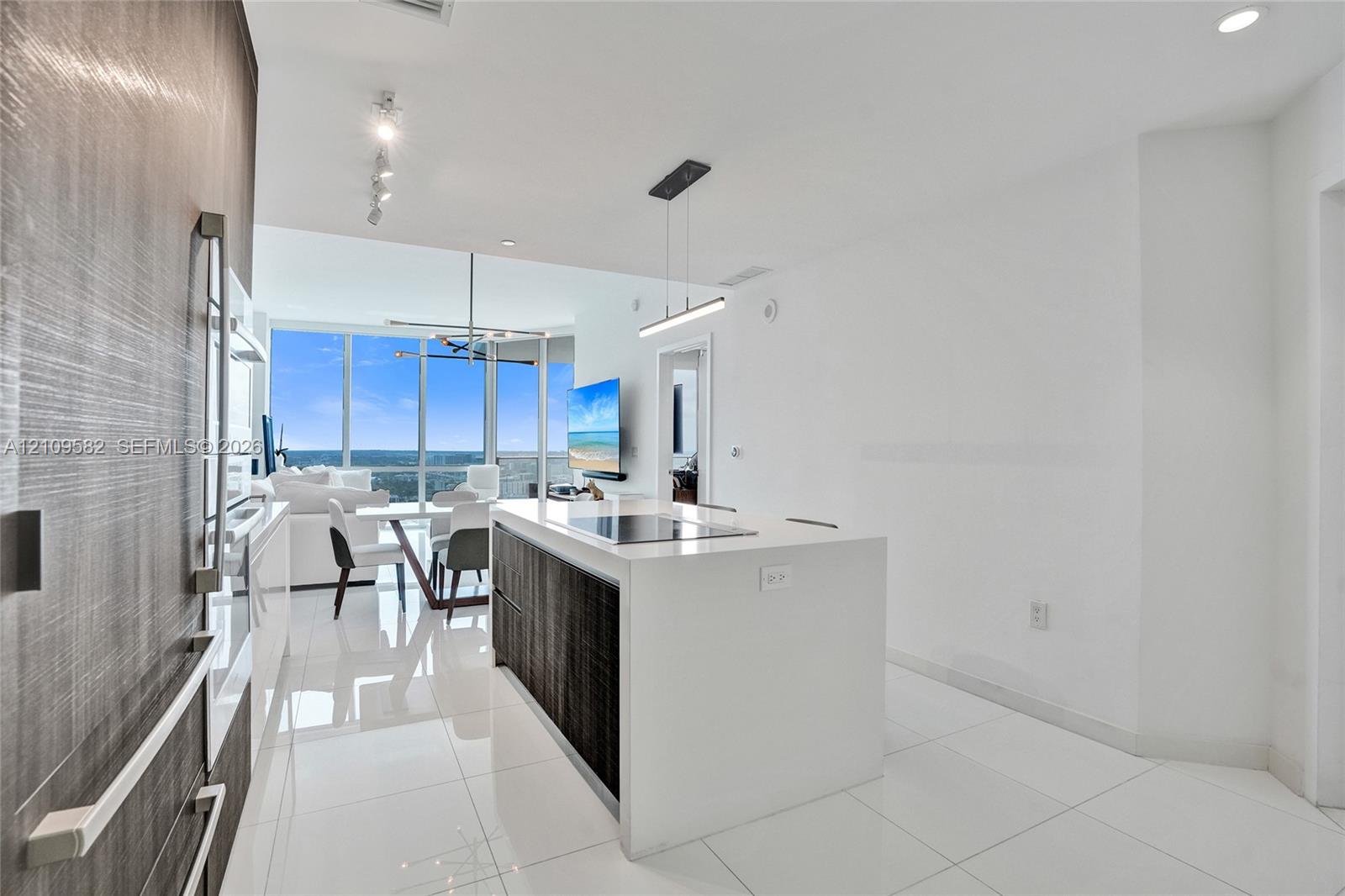 Modern kitchen featuring a central island with a solid-surface countertop and integrated cooktop, textured dark cabinetry, white tile flooring, recessed track lighting, and floor-to-ceiling windows
