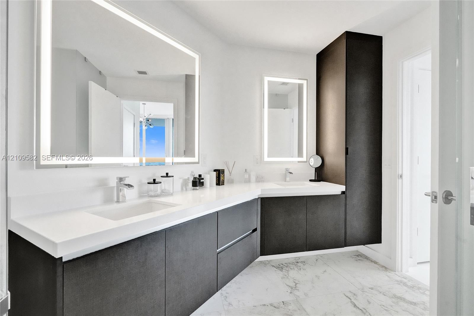 Dual-sink floating vanity with dark-finish cabinetry, solid-surface white countertops, wall-mounted illuminated mirrors, and light-tone stone flooring