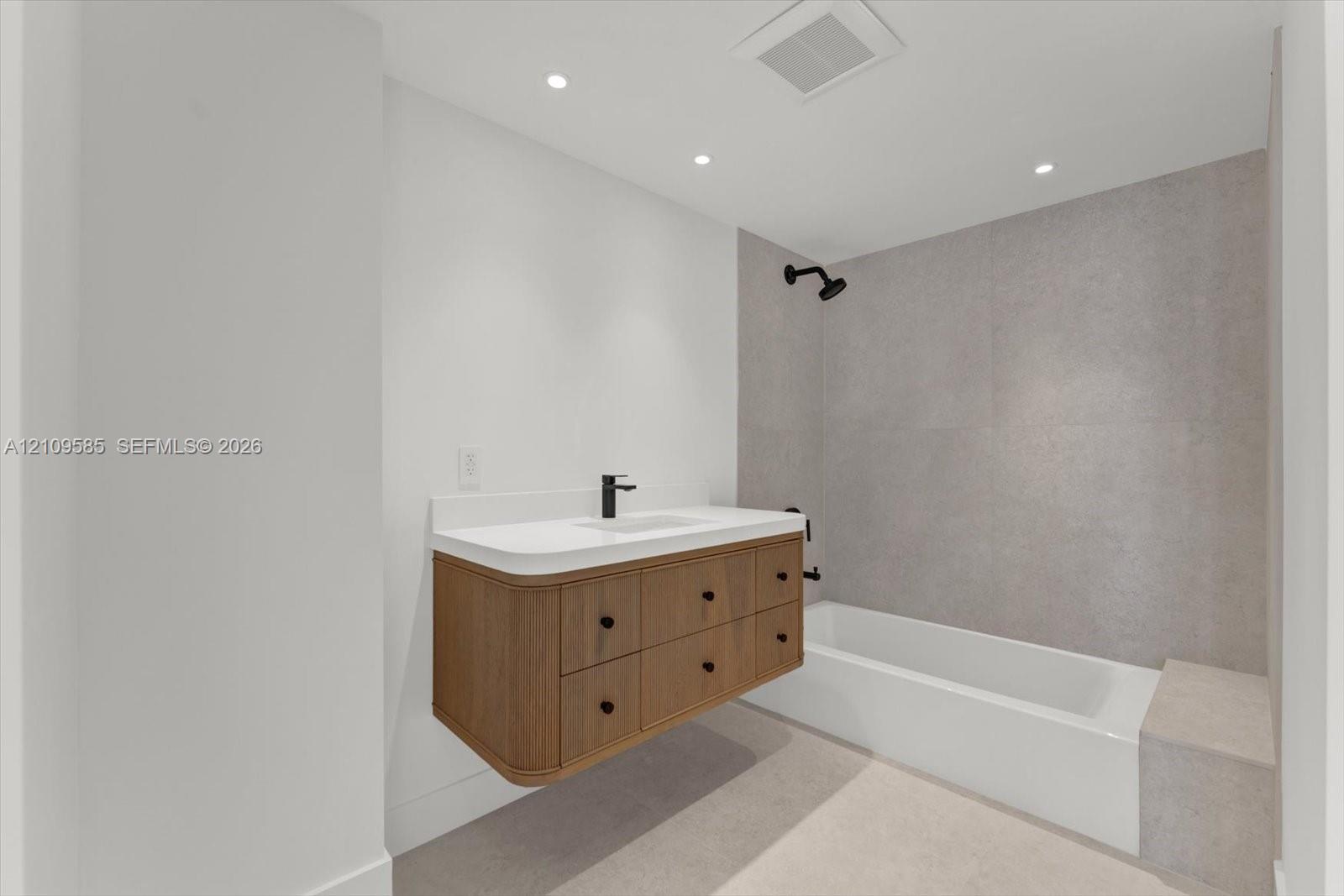 Floating wood-finish vanity with a white countertop and rectangular undermount sink, matte black faucet and shower fixtures, built-in bathtub with a stone-finish tiled surround, and recessed ceiling lighting