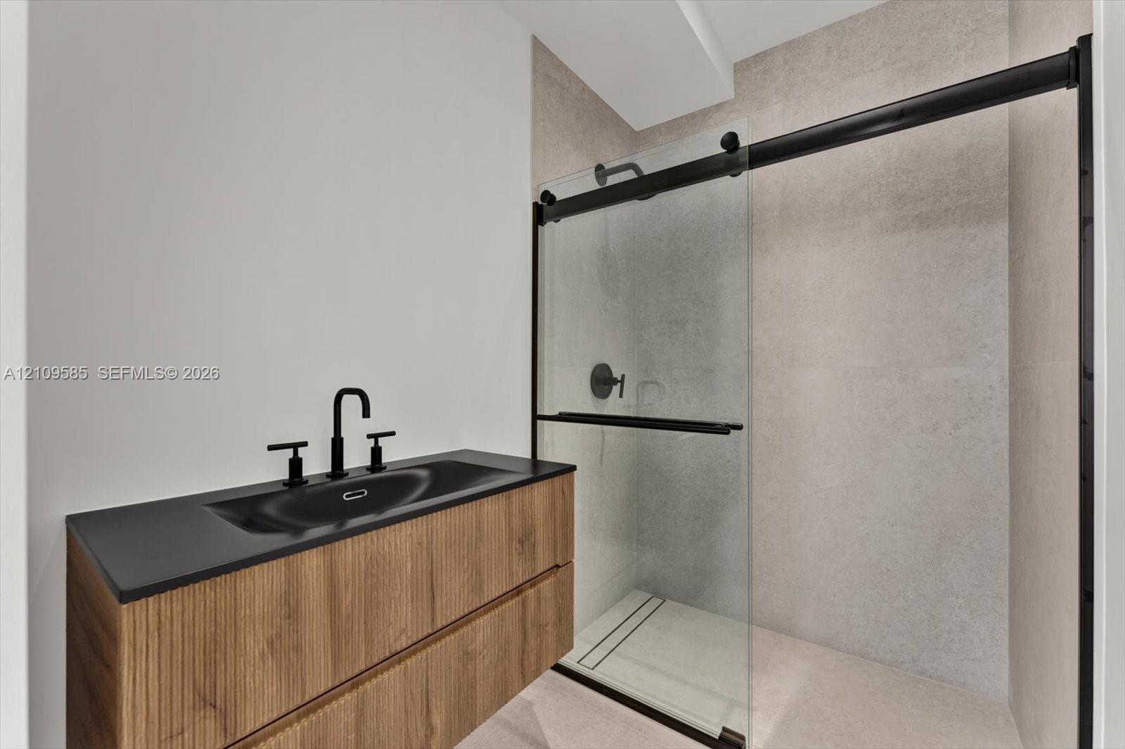 Floating wood-finish vanity with a dark countertop and integrated sink, matte black faucet, glass-enclosed shower with a sliding matte black track, and large-format wall tiling