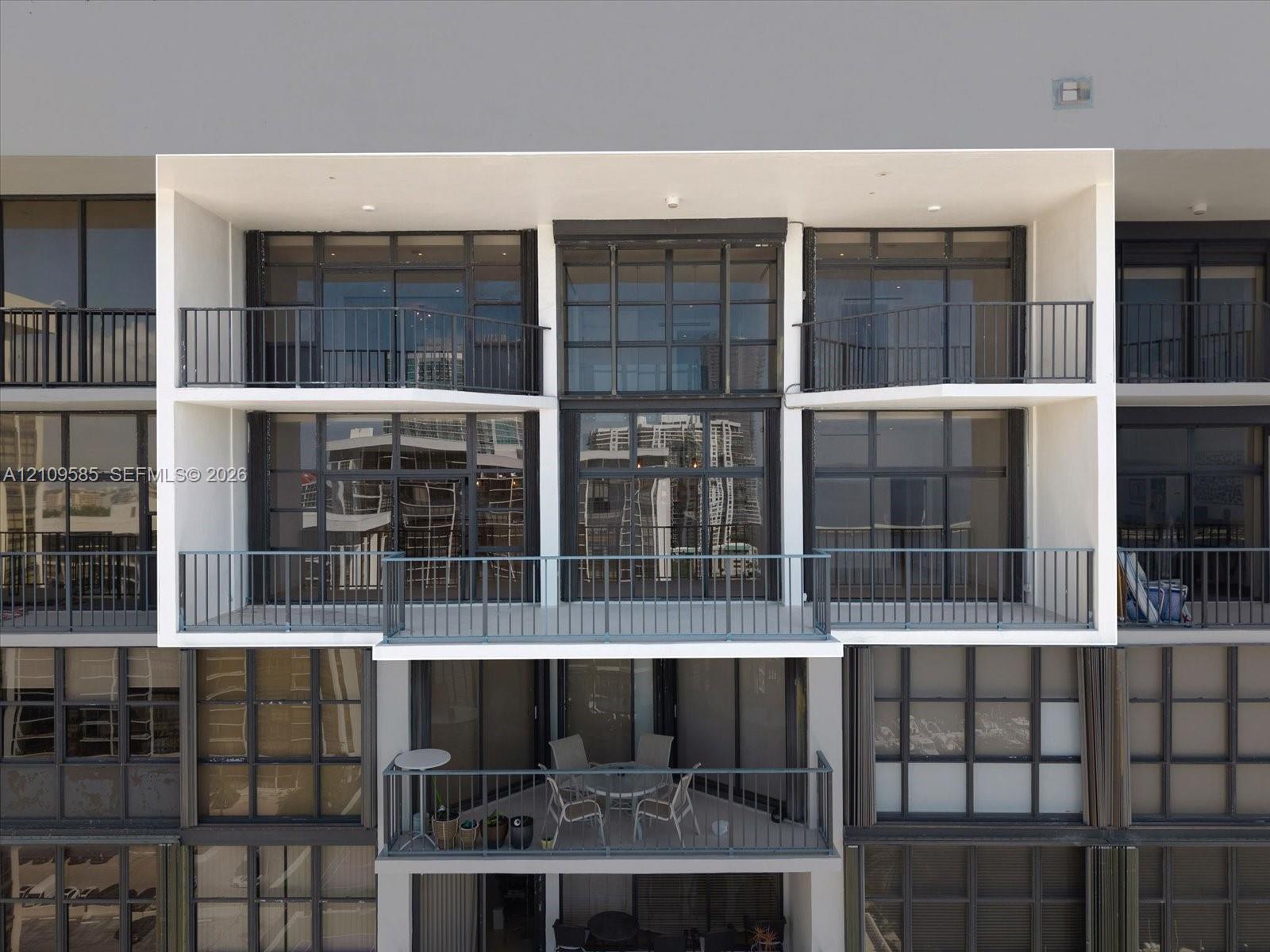 Contemporary building facade featuring multi-level open-air balconies, dark-framed floor-to-ceiling glass windows, and metal railings