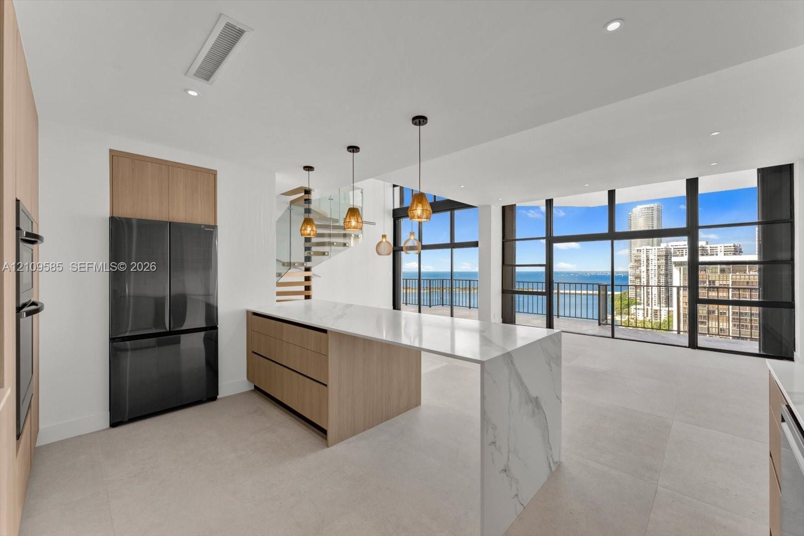 Kitchen featuring a waterfall-edge island with stone-finish countertops, flat-panel wood-finish cabinetry, floor-to-ceiling glass paneling, and pendant lighting