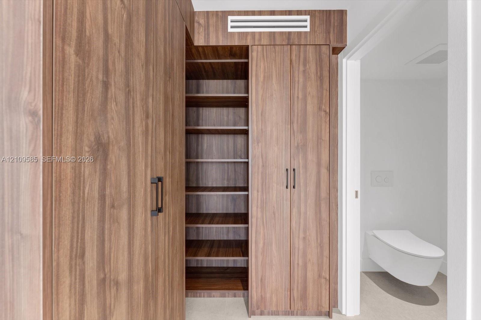 Floor-to-ceiling wood-finish cabinetry featuring open shelving, double-door storage, matte black hardware, and a wall-mounted toilet