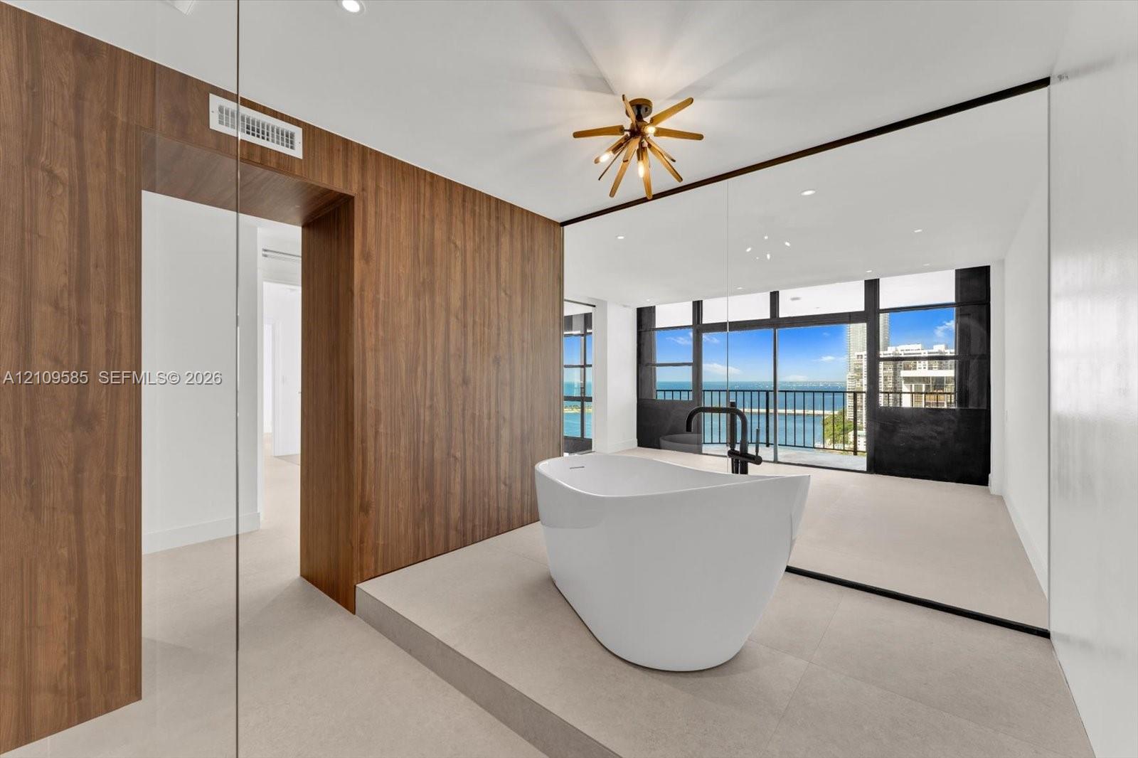 Freestanding soaking tub with matte black faucet fixture, elevated platform, wood-finish wall paneling, multi-light geometric chandelier, and floor-to-ceiling windows