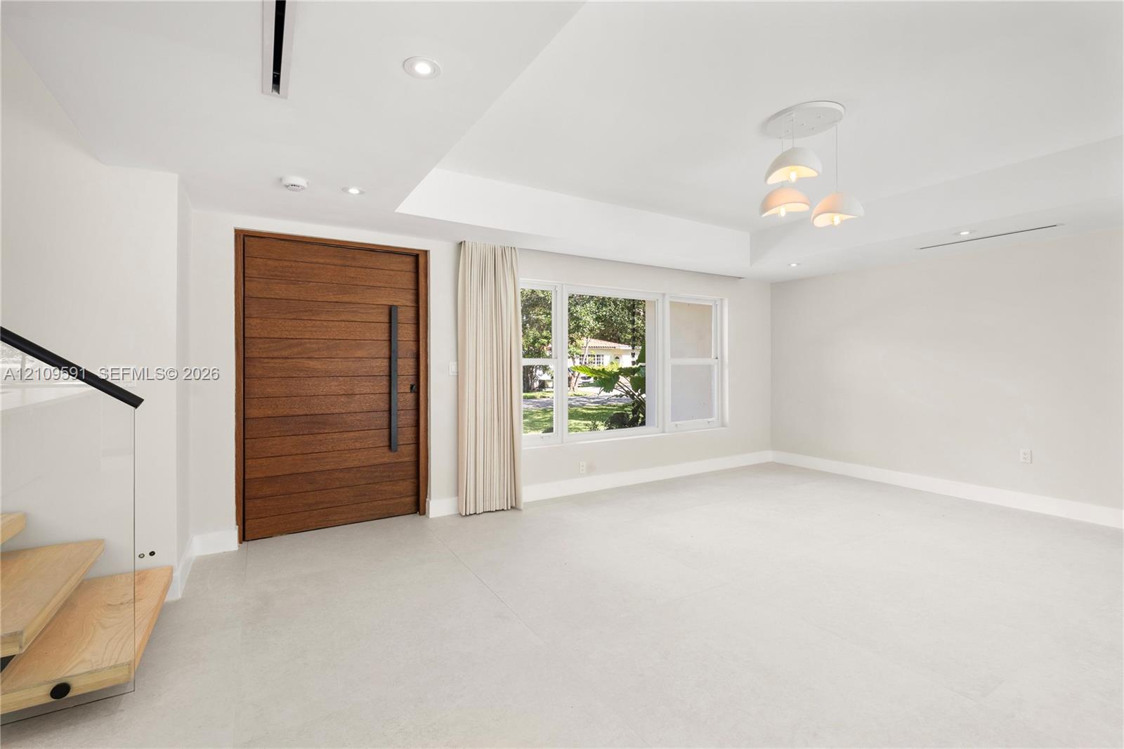 Horizontal wood-plank entry door with a vertical matte black pull handle, recessed ceiling lighting, three-globe pendant fixture, and glass-panel staircase railing