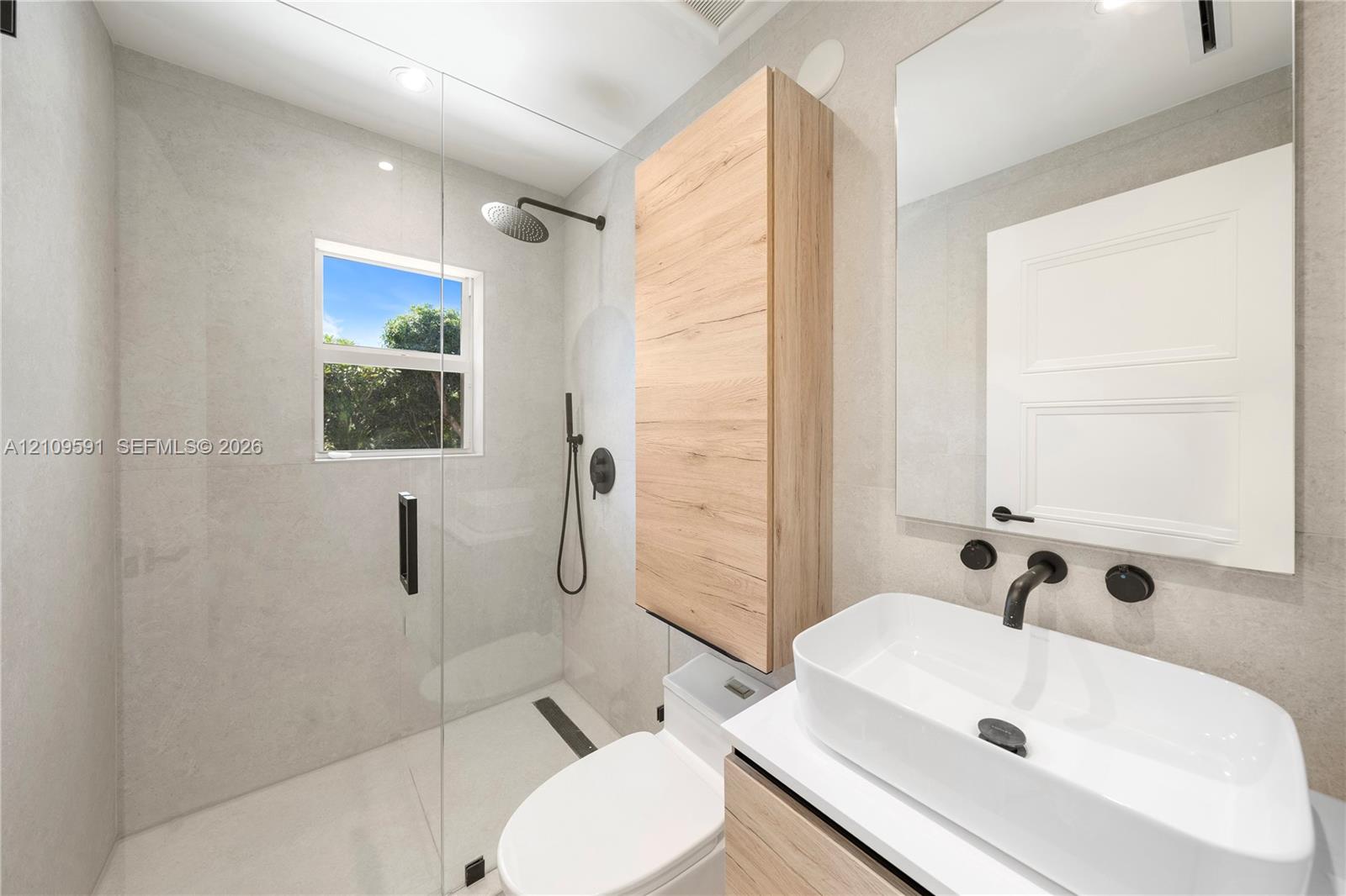 Walk-in shower with a glass enclosure and wall-mounted matte black rainfall shower head, wood-finish wall-mounted cabinetry, wall-mounted rectangular vessel sink with matte black hardware, and large-format neutral wall tile