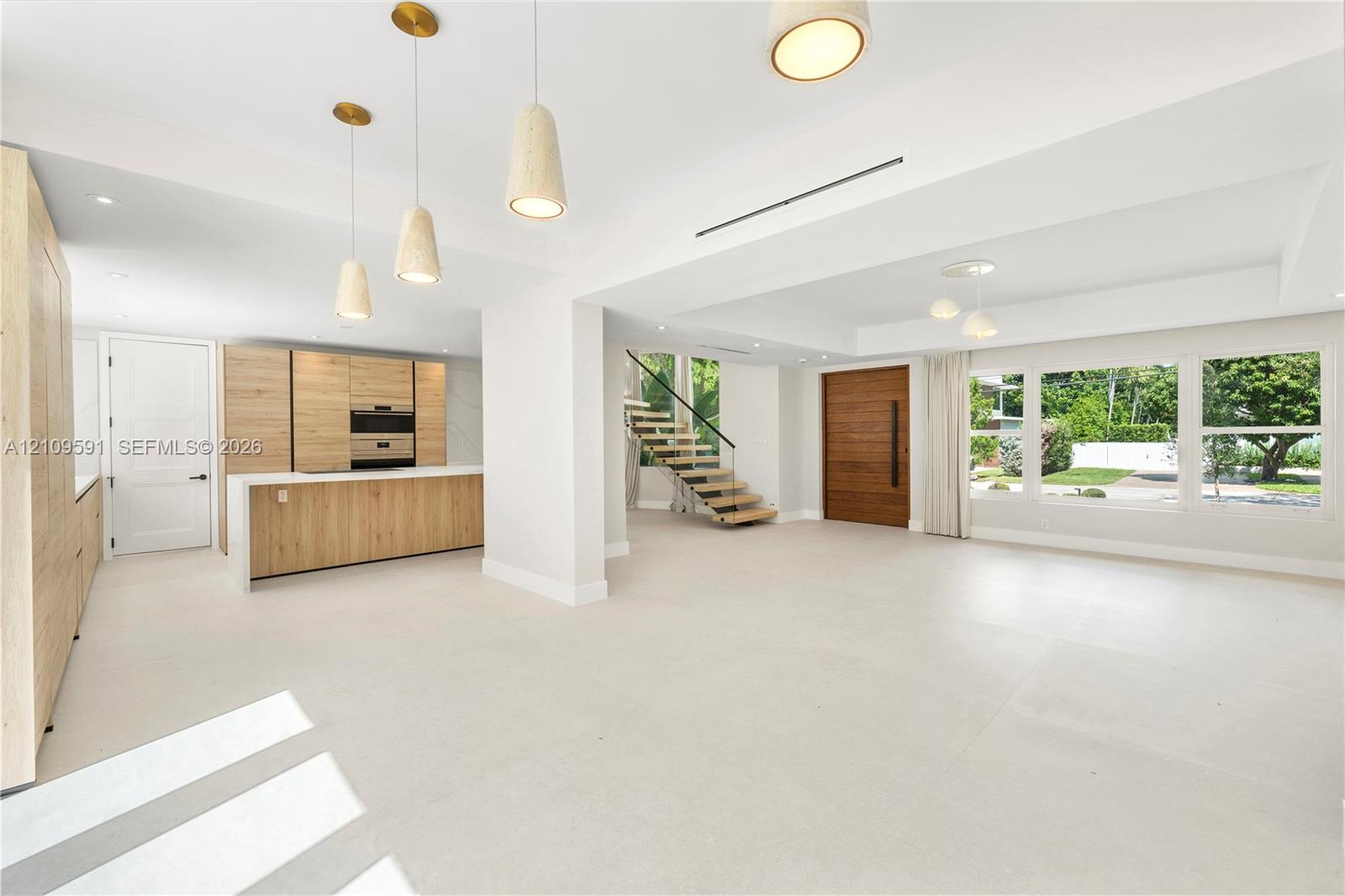 Open-concept interior featuring wood-finish cabinetry, a central kitchen island, large-format tile flooring, a floating staircase with glass railing, and a floor-to-ceiling solid wood entry door