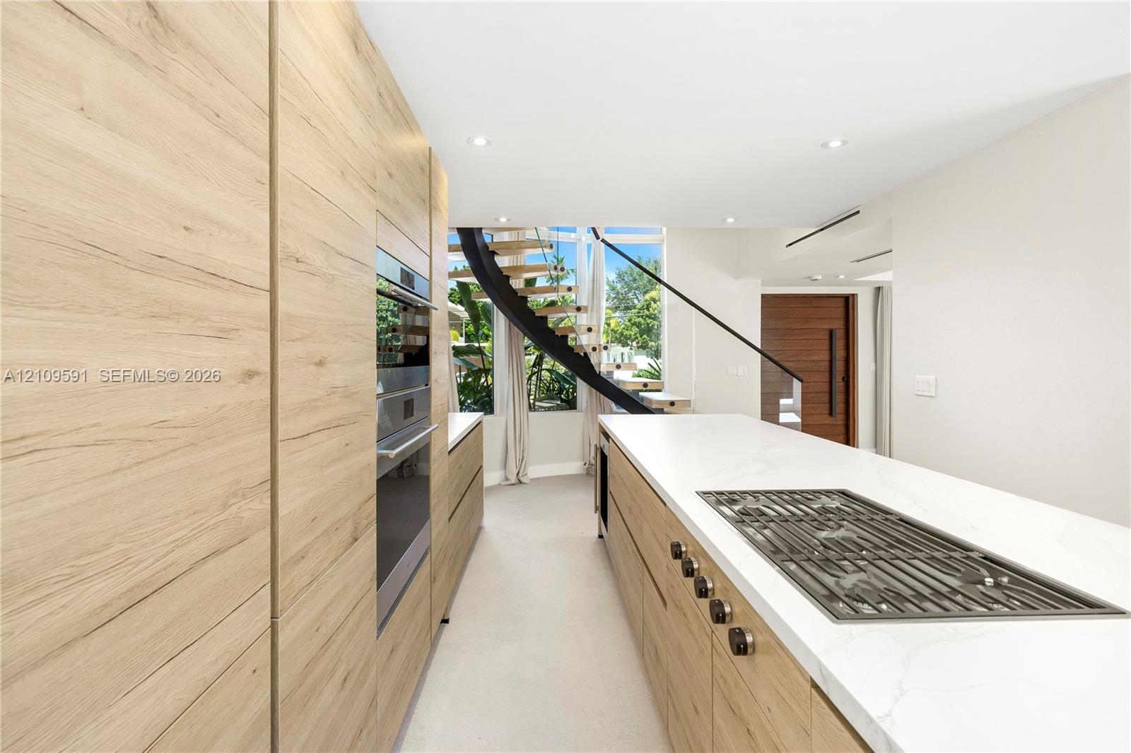 Kitchen featuring full-height wood-finish cabinetry, integrated wall oven, large island with quartz-style countertop, inset gas cooktop, and a curved staircase with floating wood treads