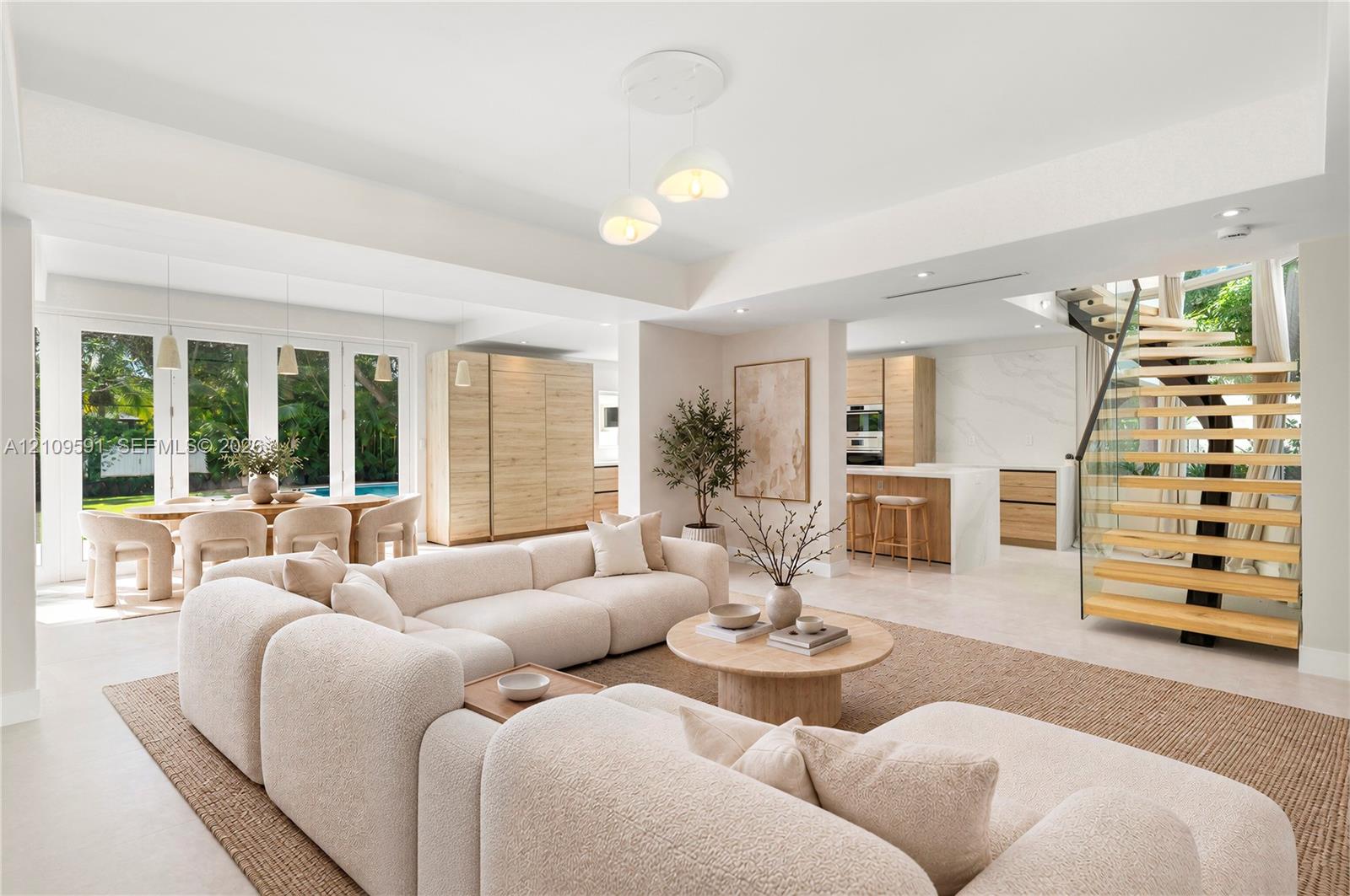 Virtually Staged: Open-concept living space featuring high tray ceilings, wood-finish cabinetry, a modern staircase with glass railings and wood treads, large-format tile flooring, and multiple glass-paneled exterior doors
