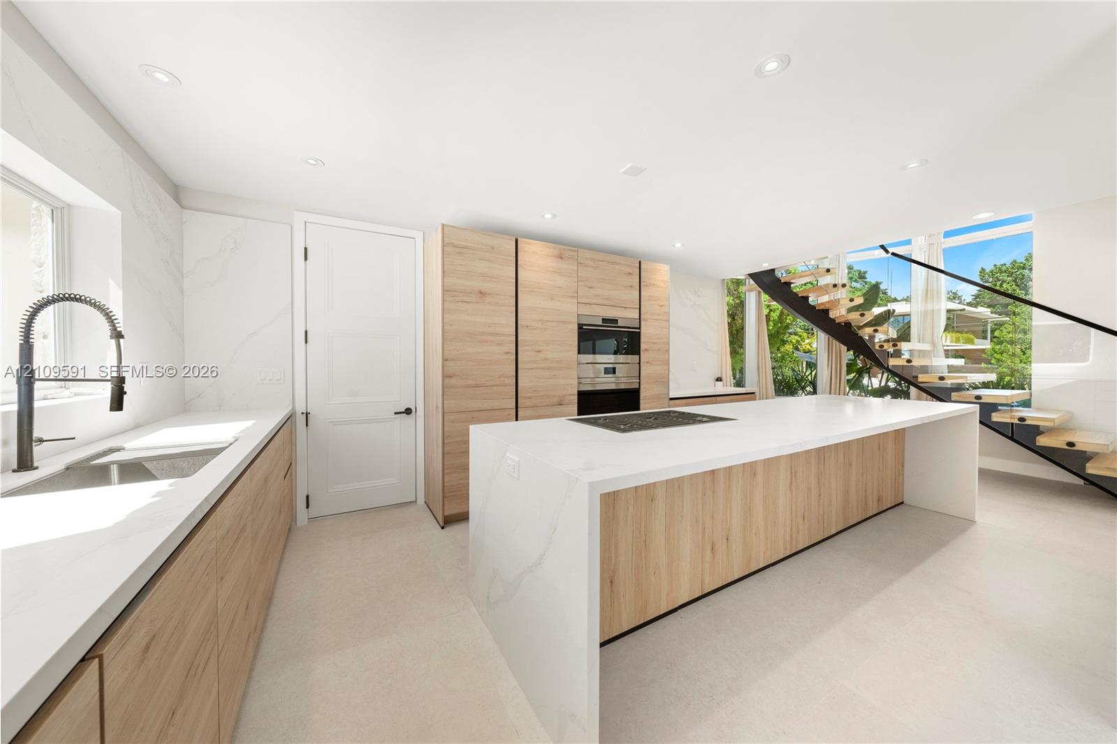 Modern kitchen featuring a waterfall-edge island, light wood-finish cabinetry, stone-style countertops, recessed lighting, and a floating staircase with black metal railings