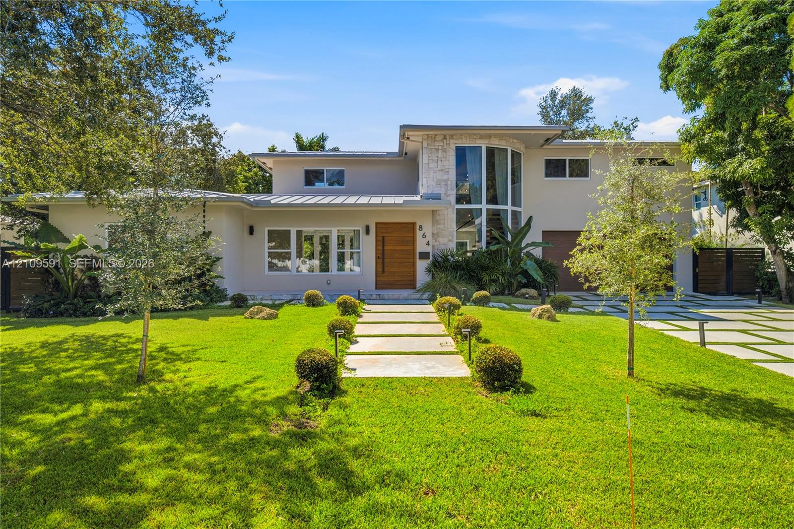 Modern exterior featuring a metal roof, stone facade accents, expansive floor-to-ceiling windows, a wood-finish front door, and a geometric concrete paver driveway