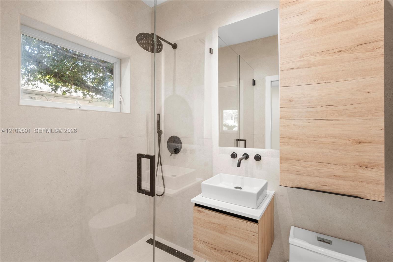 Modern bathroom featuring large-format wall tiling, a floating wood-finish vanity with a rectangular vessel sink, matte black hardware, and a glass-enclosed shower with a rainfall showerhead