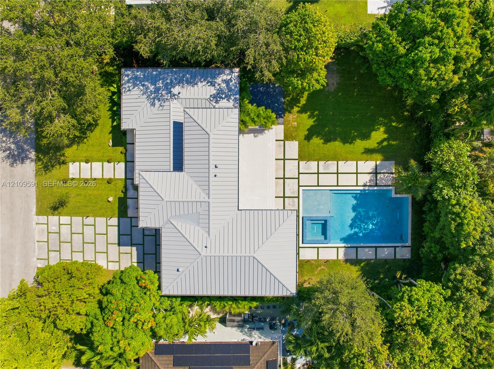 Aerial perspective of a property featuring a standing seam metal roof, geometric concrete paver hardscaping, an in-ground swimming pool with an integrated spa, and mature perimeter landscaping