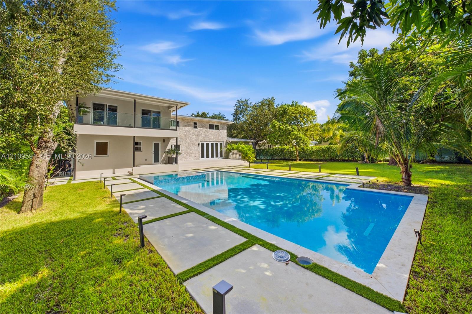 Backyard featuring a rectangular swimming pool, concrete paver decking, manicured lawn, stone-veneer exterior finish, and a second-level balcony with a glass railing