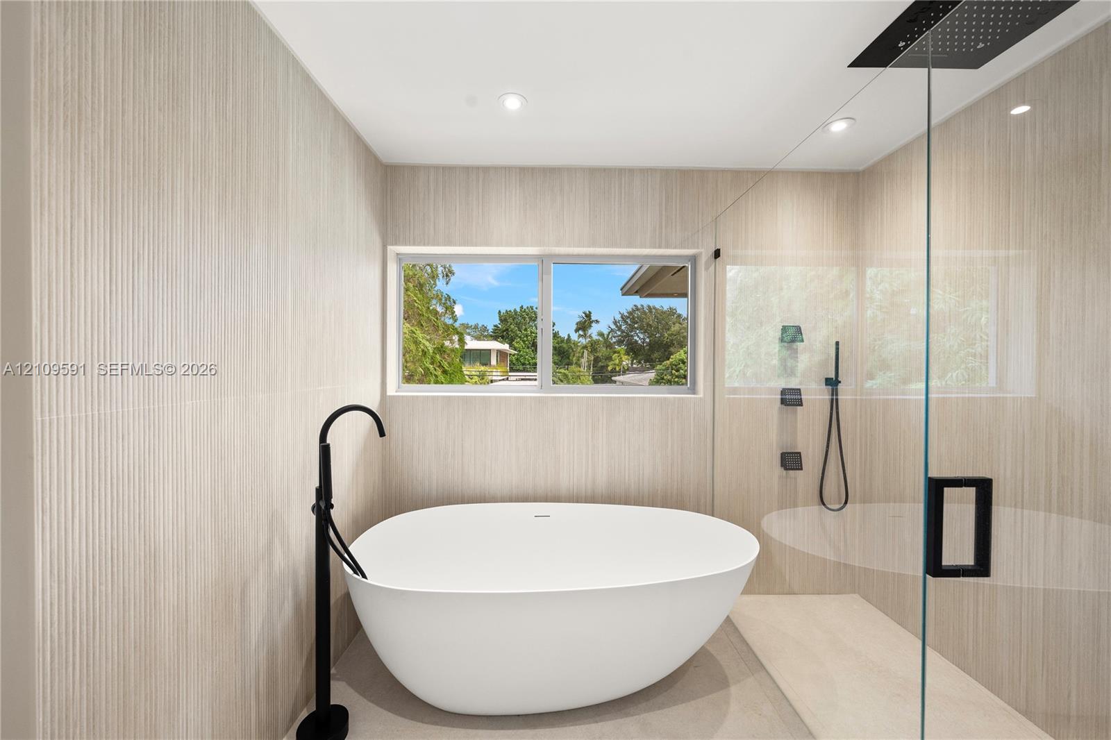 Freestanding soaking tub with matte black floor-mounted faucet, walk-in shower with glass enclosure, rain shower head, and floor-to-ceiling textured tile walls