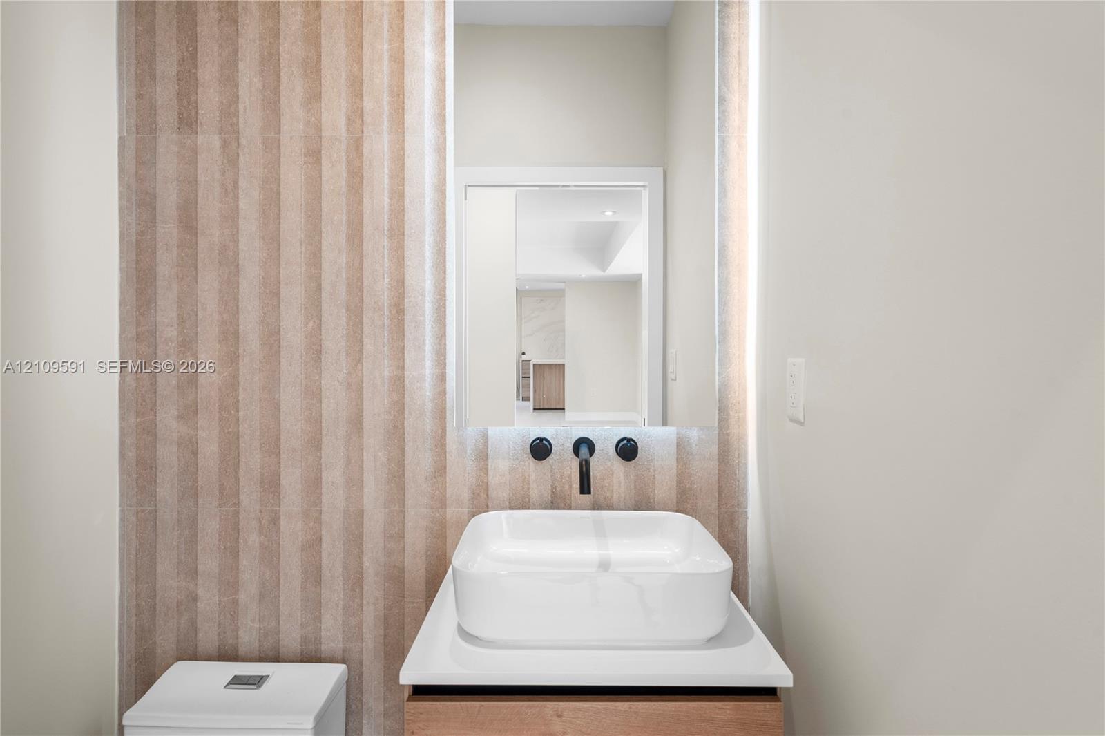Half Bath off living room. Wall-mounted matte black faucet, white rectangular vessel sink, white countertop, wood-finish vanity, and vertical textured stone wall tile