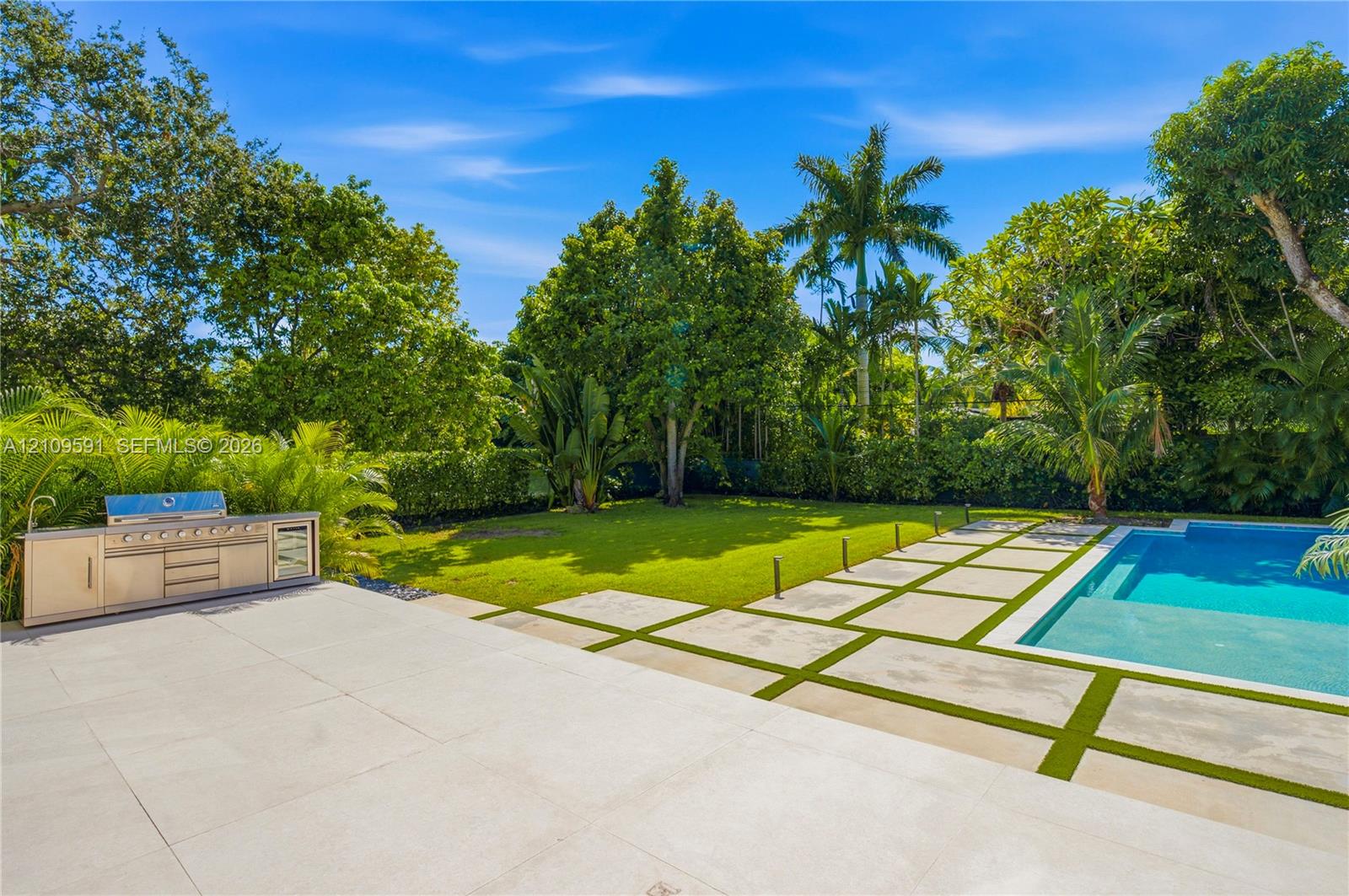 Backyard featuring a swimming pool, integrated stainless steel outdoor kitchen, stone paver patio with turf inlays, and mature landscaping