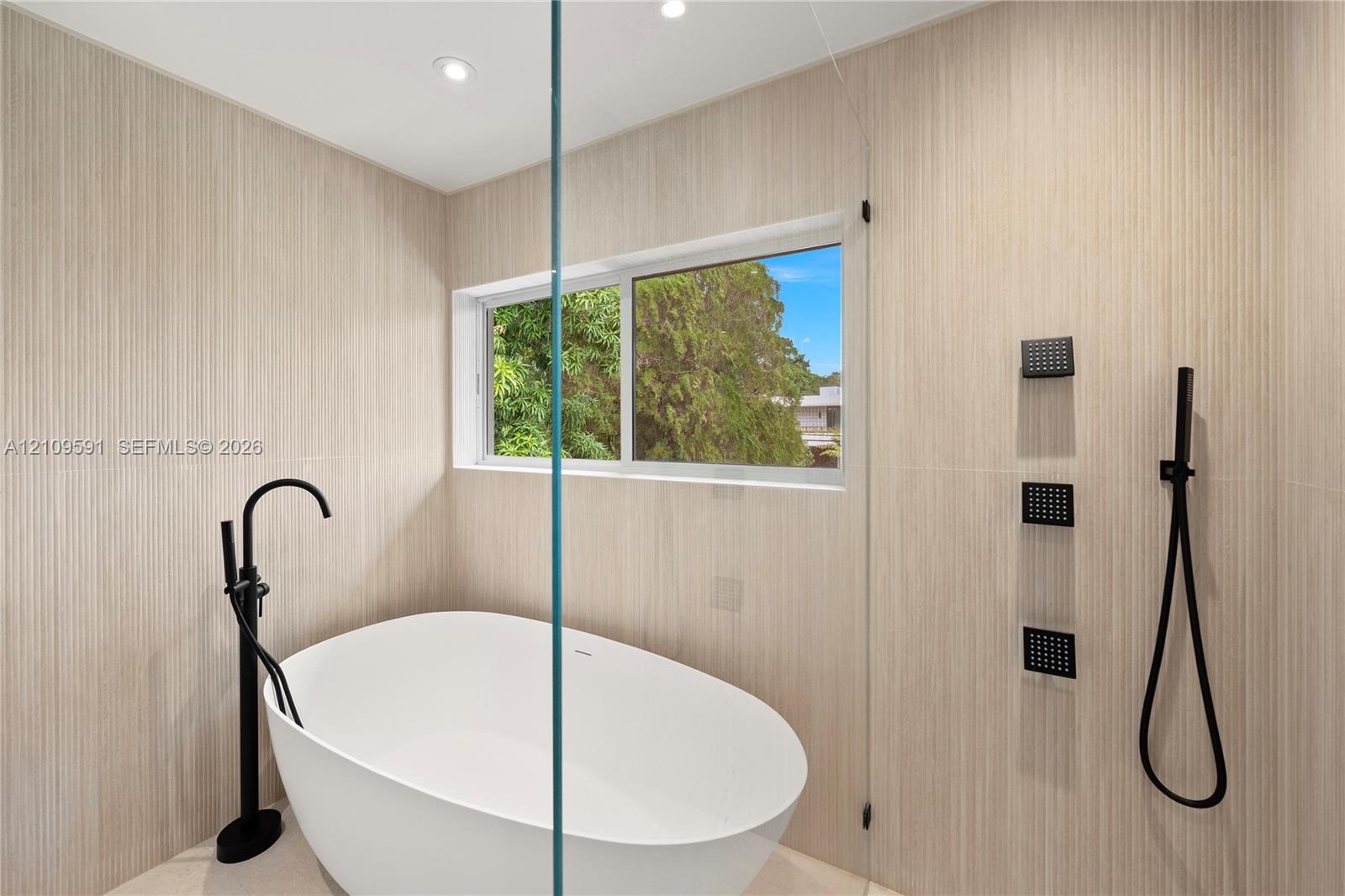 Freestanding white soaking tub with matte black floor-mounted faucet, floor-to-ceiling textured tile wall, frameless glass shower partition, and matte black rainfall shower system with handheld sprayer