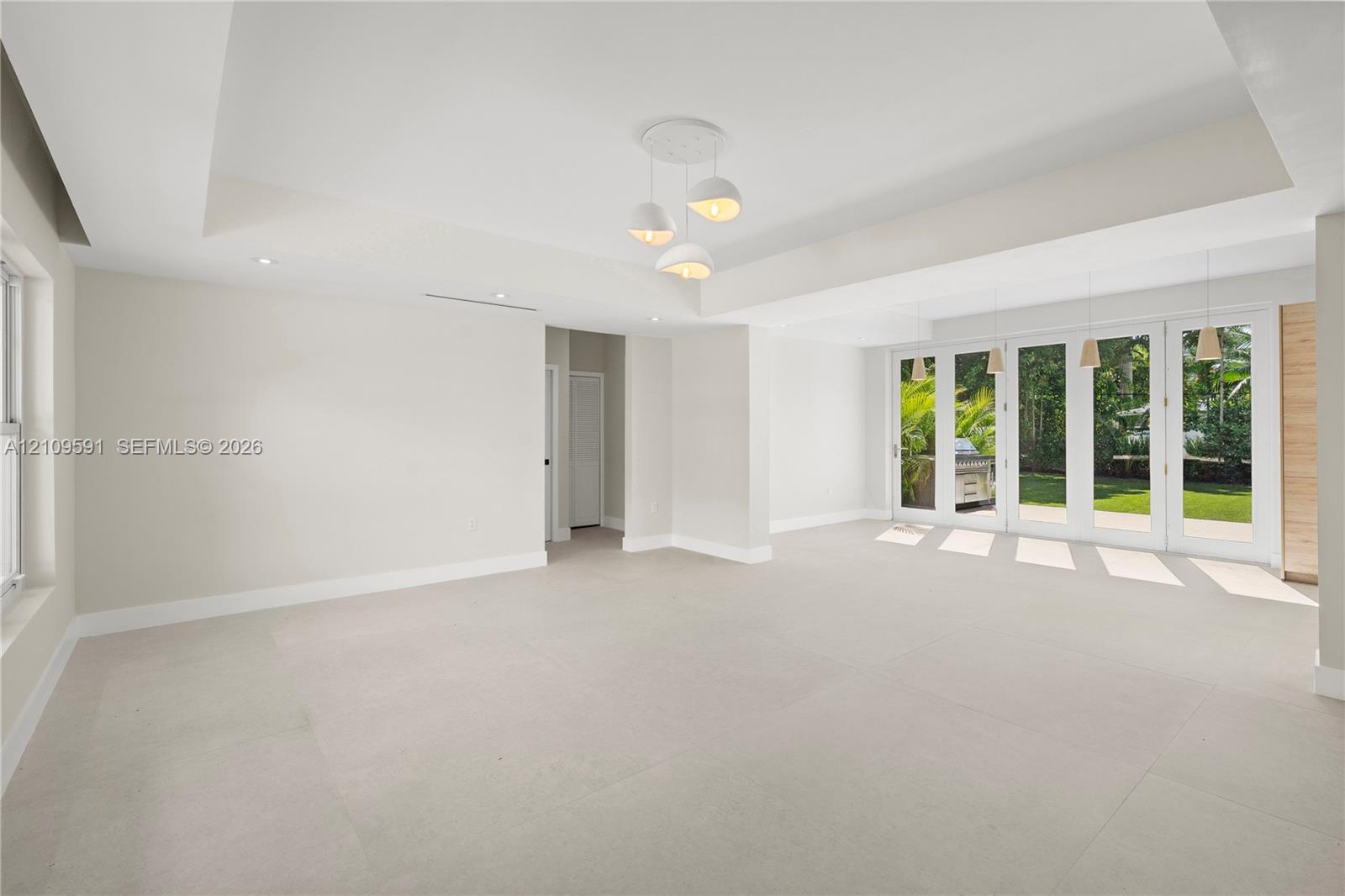 Open-concept interior featuring a recessed tray ceiling, large-format tile flooring, multi-pane glass doors, and contemporary tiered pendant lighting