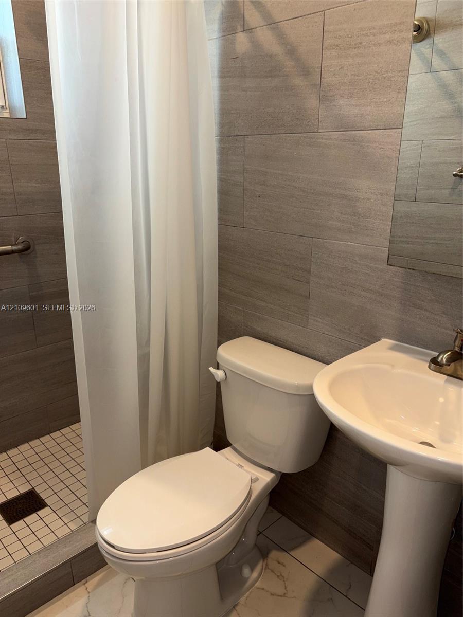 Bathroom featuring large-format neutral wall tile, white pedestal sink with metal faucet, walk-in shower with mosaic tile flooring, and wall-mounted grab bar