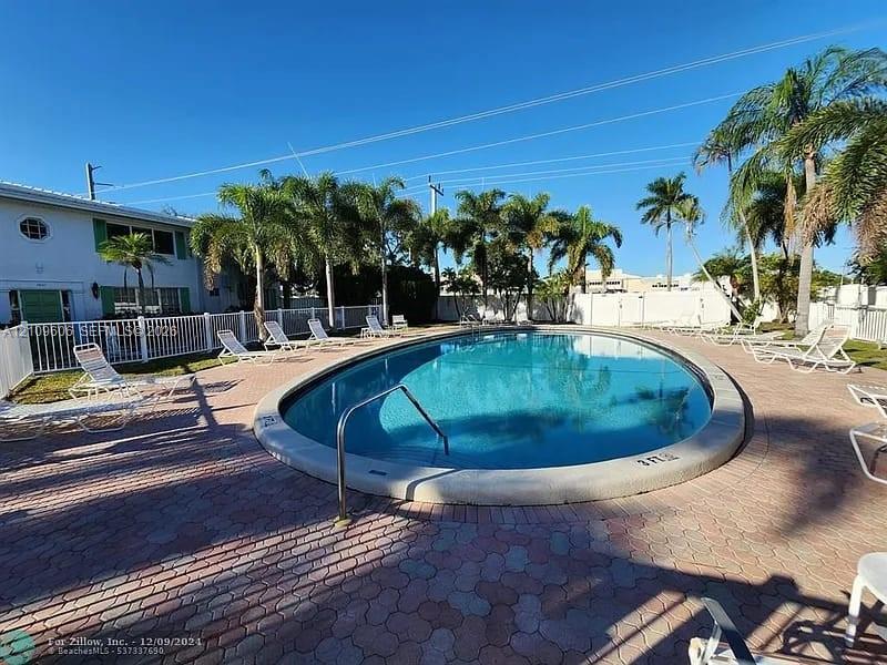 In-ground swimming pool with a concrete coping and metal handrail, surrounded by a paved patio and white perimeter fencing, accented by mature palm trees