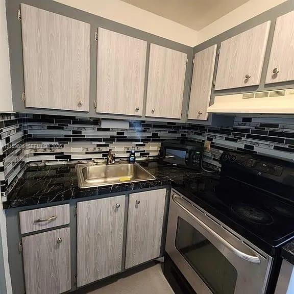 Kitchen featuring light wood-finish cabinetry, dark stone-pattern countertops, and a multi-toned horizontal tile backsplash with a stainless steel drop-in sink and built-in electric range