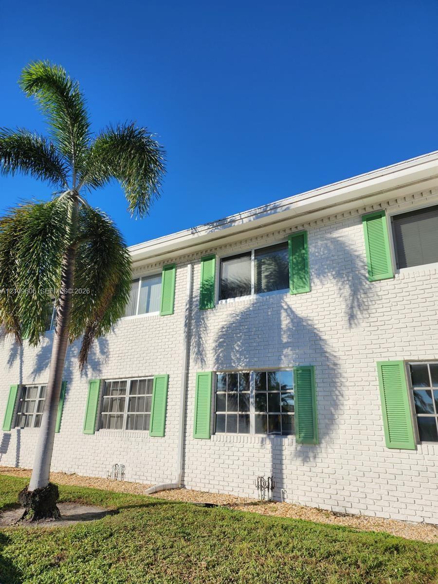 Exterior facade featuring white brick masonry, green louvered shutters, sliding windows, and a mature palm tree