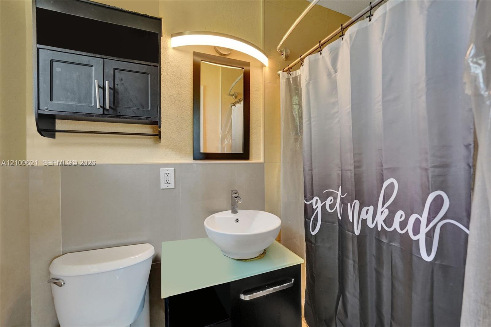 Bathroom featuring a vessel sink with a chrome faucet, a glass-top vanity, wall-mounted cabinetry, a curved vanity light fixture, and tile wall surfacing