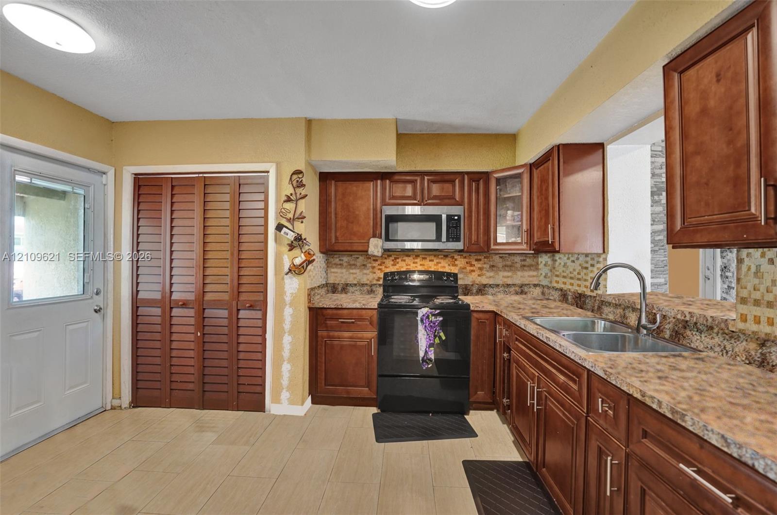 Kitchen featuring wood cabinetry, stone-pattern laminate countertops, tiled backsplash, stainless steel microwave, and dual-basin stainless steel sink