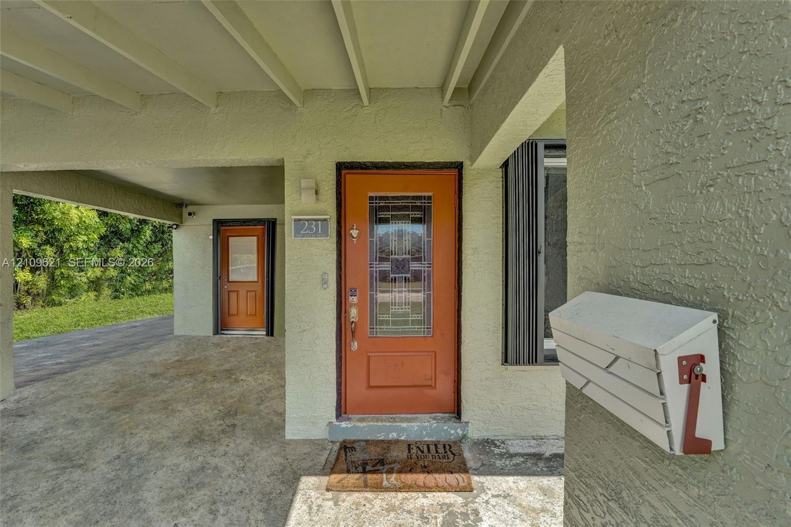 Covered entryway featuring a textured exterior finish, two distinct orange doors with decorative glass inserts, and a wall-mounted metal mailbox