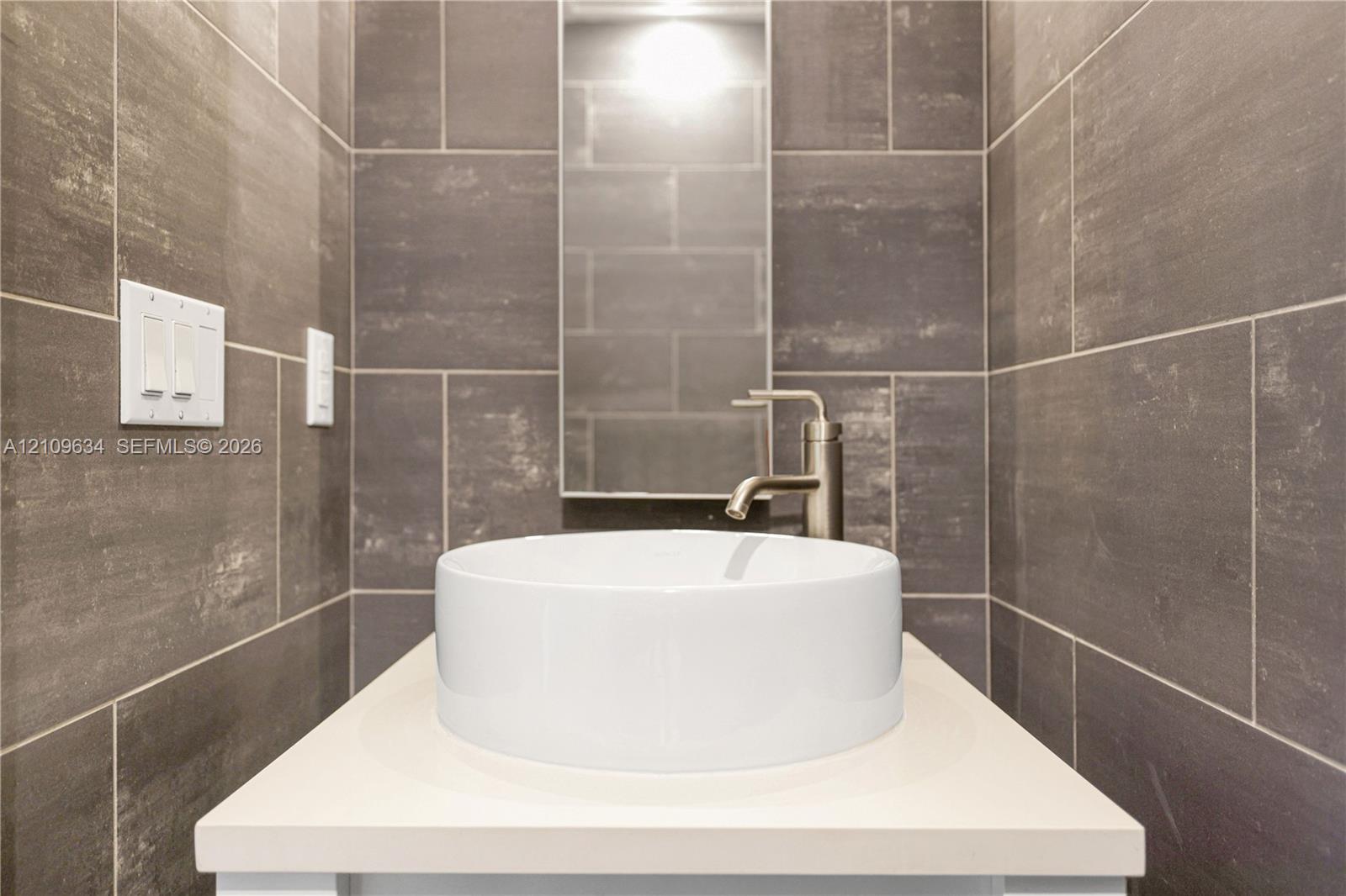 Rectangular vanity featuring a solid surface countertop, white ceramic vessel sink, brushed-finish faucet, and full-height wall tiling