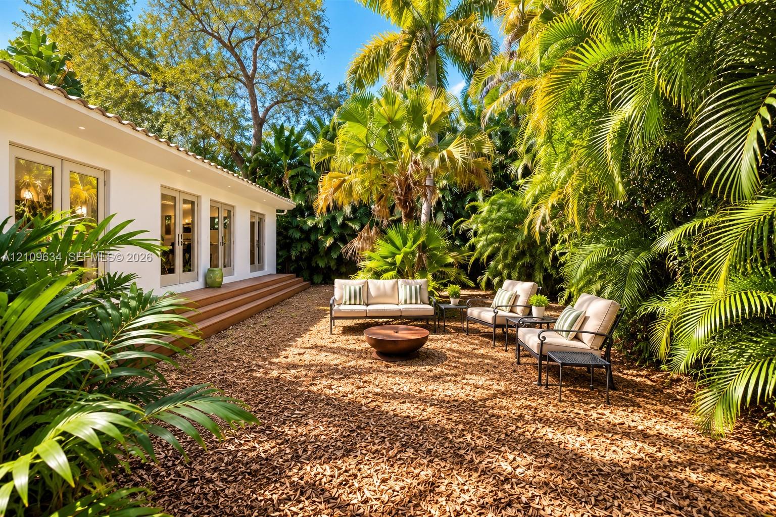 Exterior patio area featuring a low-profile wood-finish deck, multi-panel glass French doors, and mature tropical landscaping