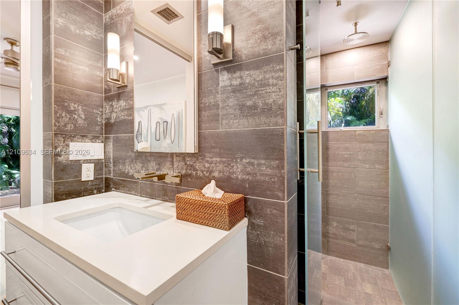 Wall-mounted vanity featuring a white countertop and integrated basin, large-format grey tile wall cladding, glass-enclosed shower, rain-style showerhead, and brushed metal fixtures
