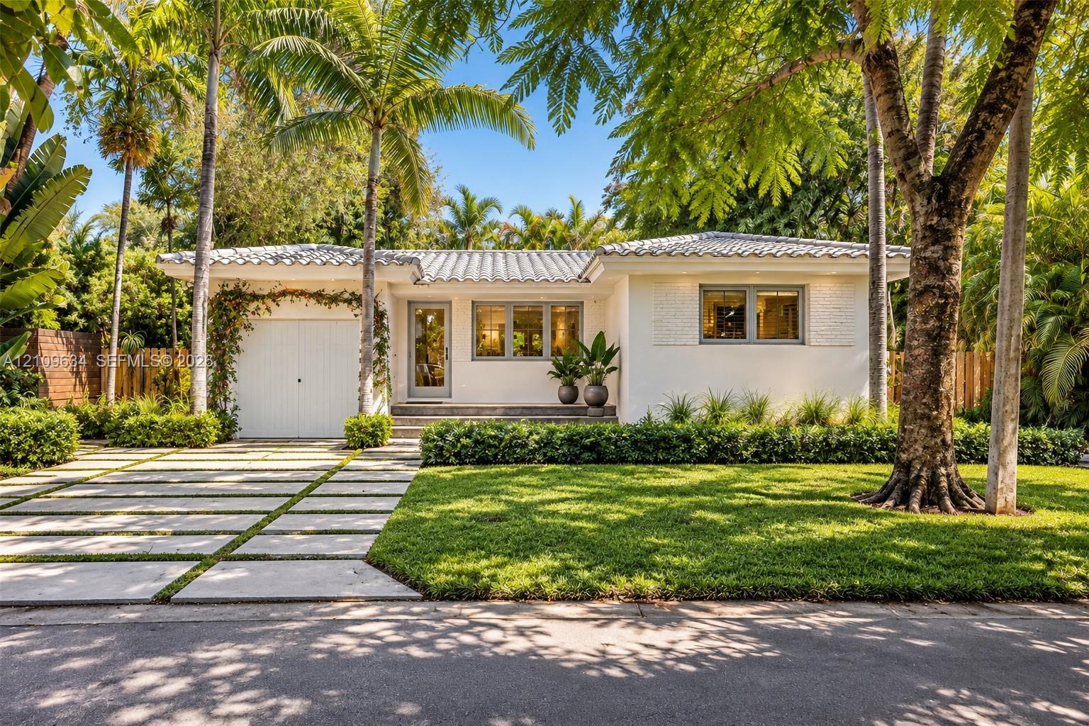 Single-story exterior featuring a light-toned stucco facade, tiered tile roofing, paver driveway with integrated turf strips, and mature palm and shade trees