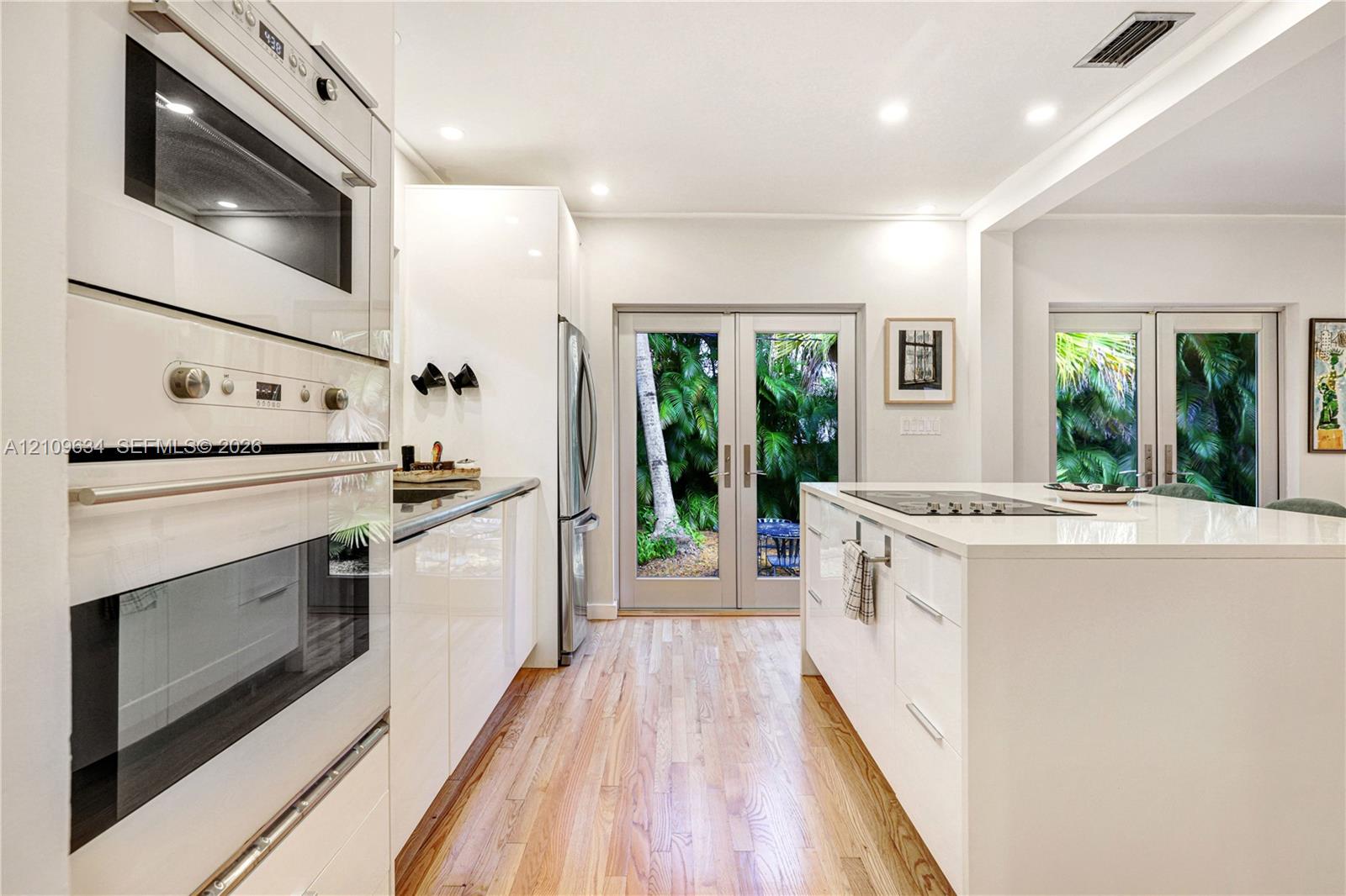 Kitchen featuring high-gloss white cabinetry, wood-finish flooring, a central island with a cooktop, built-in wall oven and microwave, and glass-paneled double doors