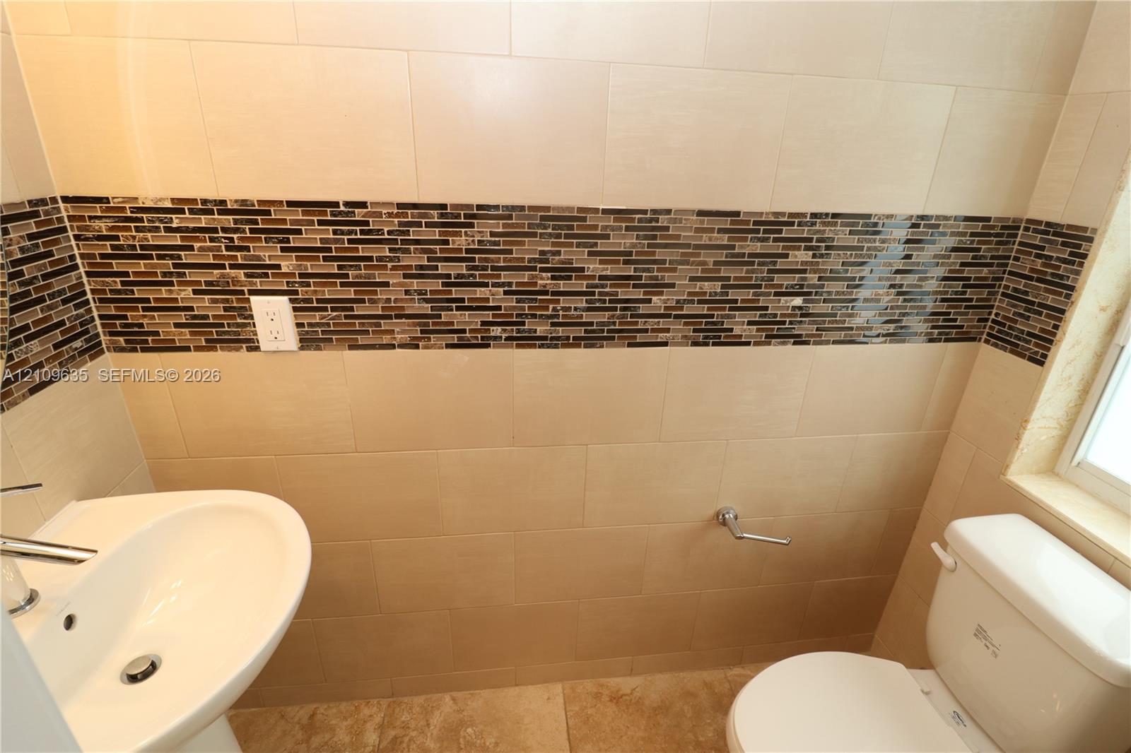 Pedestal sink with chrome faucet, porcelain tile flooring, and wall surfaces featuring large-format beige tiles accented by a horizontal glass mosaic border