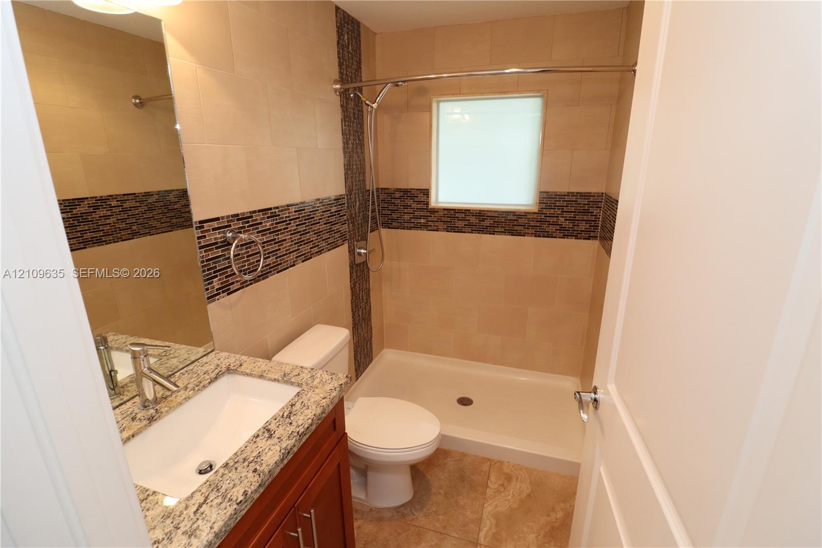 Bathroom featuring stone-finish countertop with an integrated basin, wood-finish cabinetry, walk-in shower with tile surround, mosaic decorative tile accents, and neutral floor tile