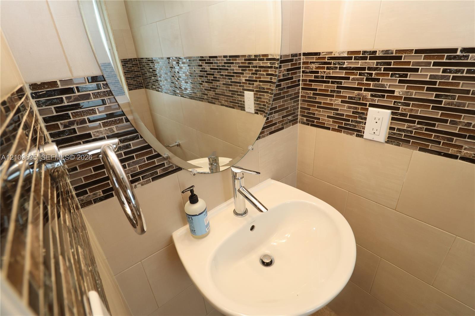 Wall-mounted white basin sink featuring a single-handle chrome faucet, oval frameless mirror, and horizontal mosaic tile backsplash with large-format neutral wall tiles