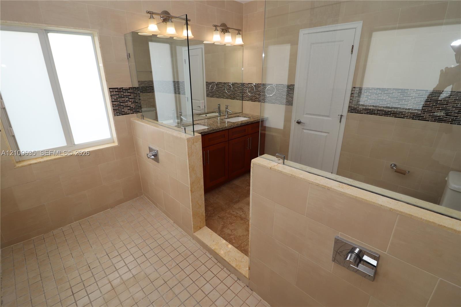 Bathroom featuring a dual-sink vanity with stone countertop, frameless glass shower enclosure, large-format tile surround with glass mosaic inlay, and tile flooring