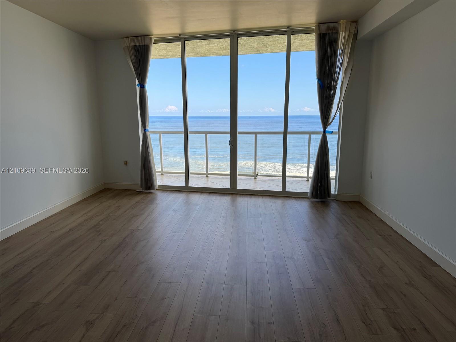 Floor-to-ceiling glass sliding doors opening to an exterior balcony with a railing, complemented by wood-finish flooring and direct ocean scenery