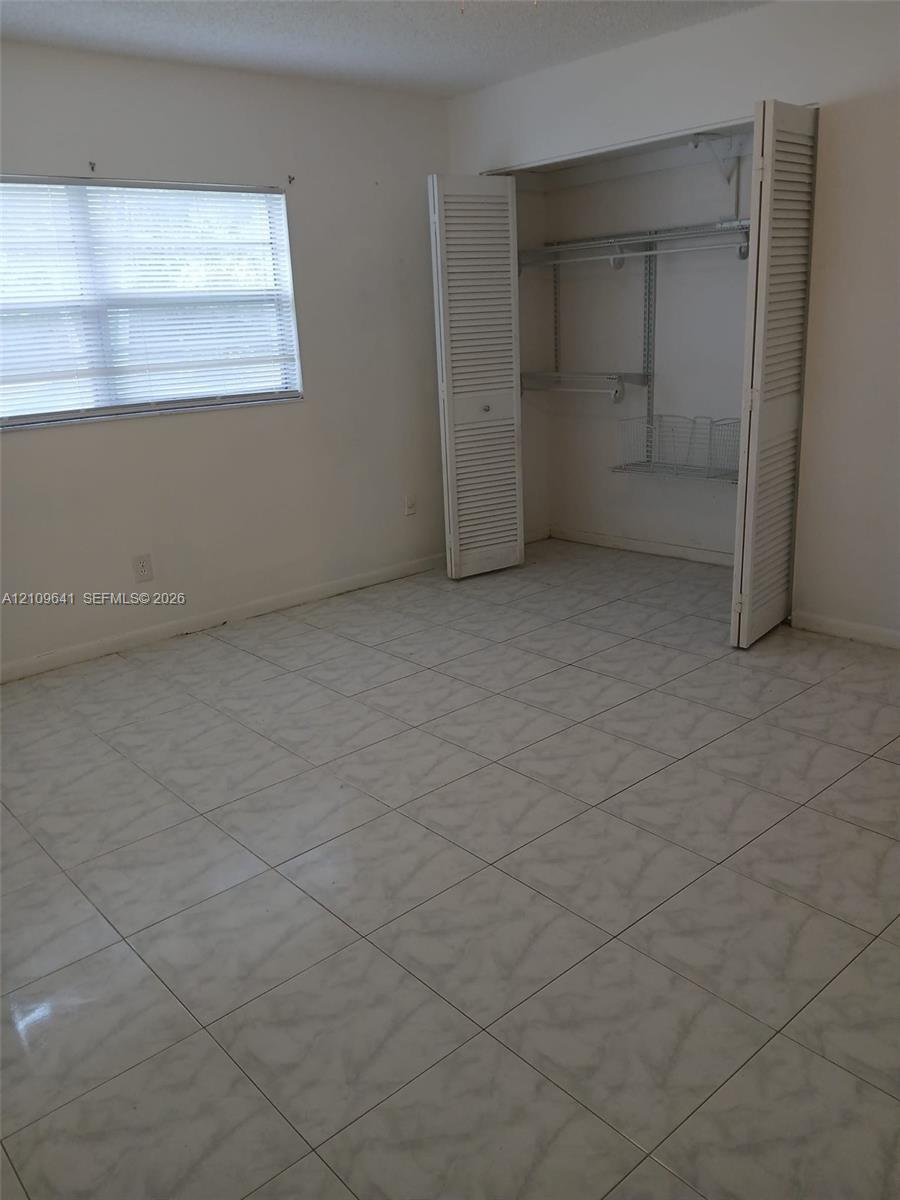 Room featuring neutral tile flooring, window with built-in blinds, and a closet with bifold louvered doors and integrated wire shelving Room featuring neutral tile flooring, window with built-in blinds, and a closet with bifold louvered doors and integrated wire shelving