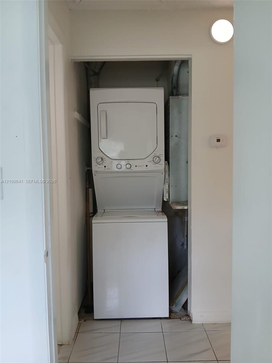 Laundry closet containing a stacked washer and dryer unit, featuring white ceramic tile flooring and a wall-mounted light fixture Laundry closet containing a stacked washer and dryer unit, featuring white ceramic tile flooring and a wall-mounted light fixture