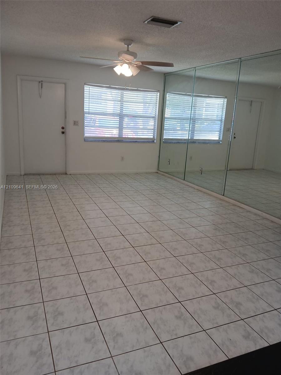Room interior featuring large-format tile flooring, wall-to-wall mirrored panels, a ceiling fan with integrated lighting, and a single-pane window Room interior featuring large-format tile flooring, wall-to-wall mirrored panels, a ceiling fan with integrated lighting, and a single-pane window