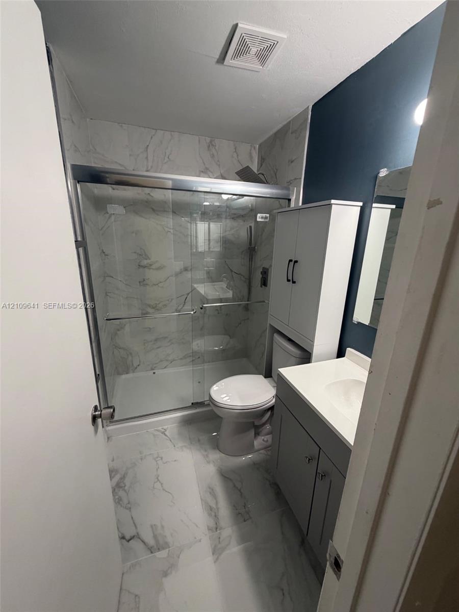Bathroom featuring large-format stone-patterned tile flooring and wall surrounds, a glass-enclosed shower with metallic hardware, a grey vanity with a solid-surface countertop, and built-in vertical cabinetry