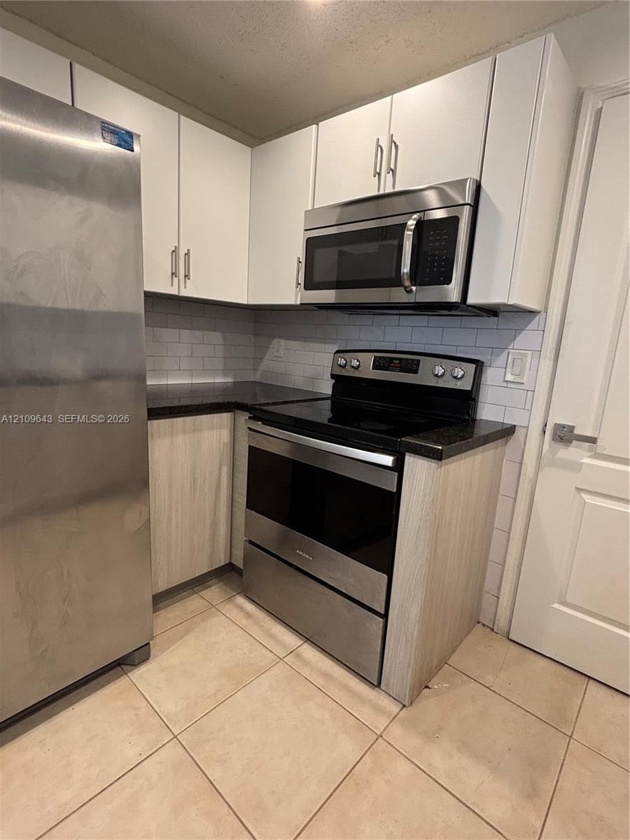 Kitchen featuring stainless steel appliances, dark stone-finish countertops, subway tile backsplash, white upper cabinetry, and wood-finish lower cabinetry