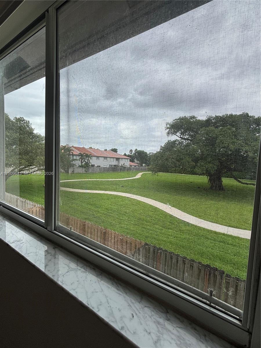 Horizontal sliding window featuring a stone-finish sill, revealing a manicured lawn with a curved paved path, mature canopy tree, and wood fencing