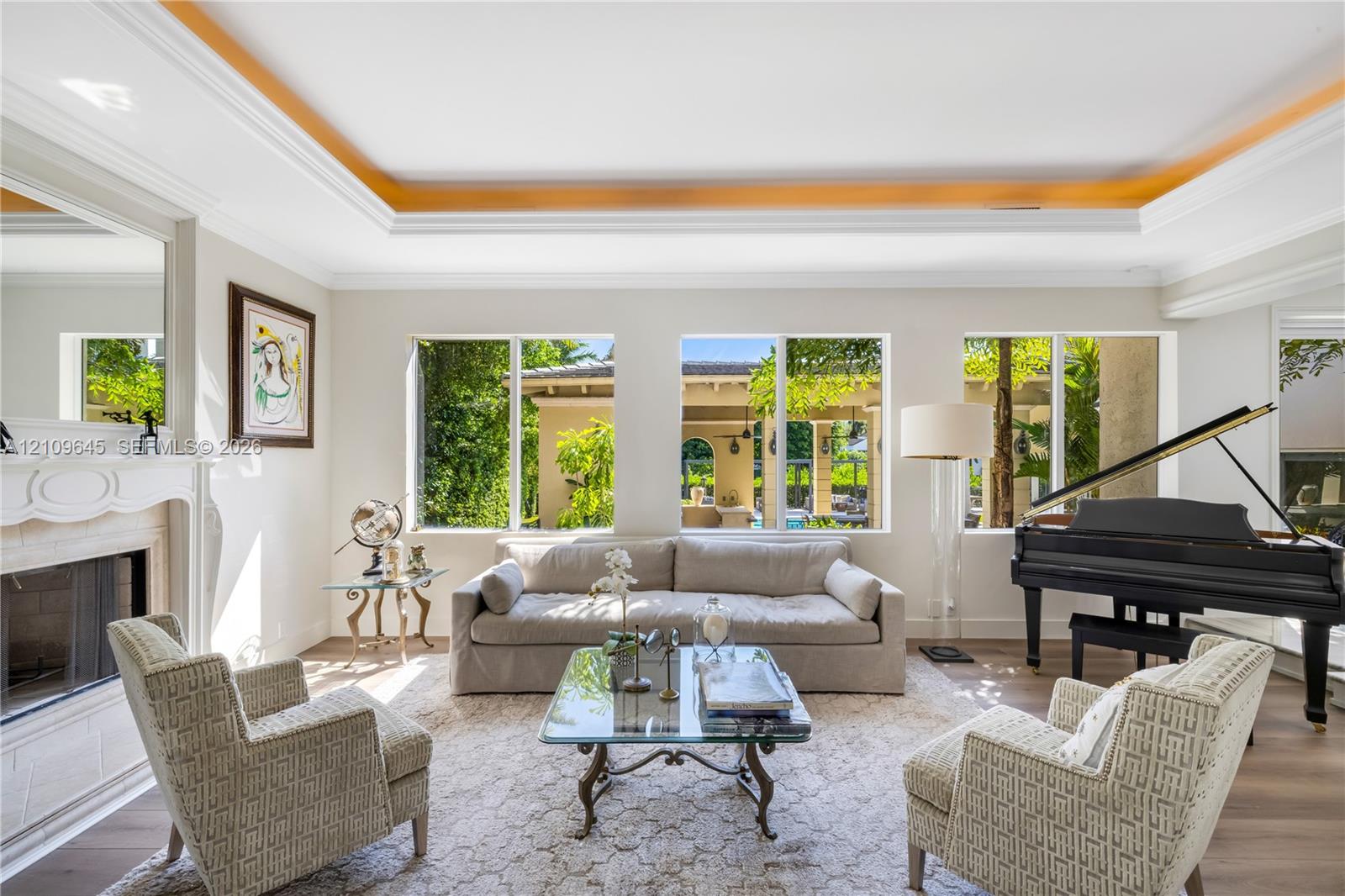 Living area featuring tray ceilings, wood-finish flooring, and a fireplace with a detailed mantel, illuminated by large windows
