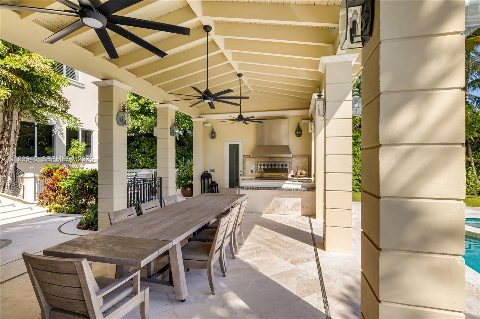 Covered outdoor kitchen and dining area featuring a stone-tile patio, built-in stainless steel range hood, stone-clad counter, multi-blade ceiling fans, and decorative wall-mounted lighting