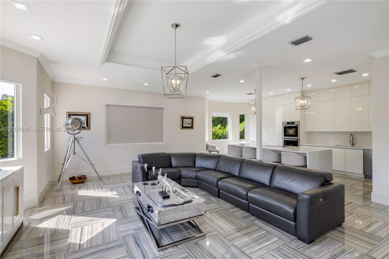 Open-concept area featuring high-gloss cabinetry, stone tile flooring, tray ceilings, recessed lighting, and a kitchen island with integrated sink